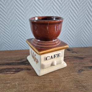 Op de afbeelding: Een bruine en witte keramische koffiemolen met het woord "CAFE" aan de voorkant. De molen is een decoratief stuk en is niet functioneel.