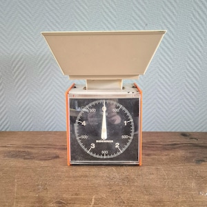 Puede incluir: Una báscula de cocina vintage naranja y negra con un cuenco blanco. La báscula tiene un dial redondo con números negros y una aguja negra. La báscula está etiquetada como "SOEHNLE" y tiene marcas para 500 gramos.