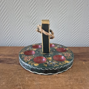 May include: A hand-painted wooden egg holder with a dark green base and floral accents. The holder has six red egg-shaped indentations and a black handle with a gold top and rope detail. The holder is on a wooden surface.