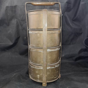 May include: A vintage brass tiffin carrier with four stacked containers and a handle. The carrier has a worn, aged patina and is made of metal.