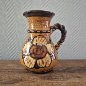 Op de afbeelding: Een kleine, bruine keramische kan met een bloemmotief. De kan heeft een gedraaide handgreep en een donkerbruine rand. Het bloemmotief is in tinten bruin en beige, met zwarte accenten. De kan staat op een houten oppervlak.