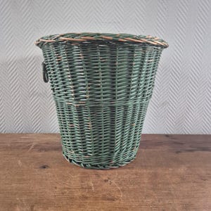 Könnte beinhalten: Ein konisch geformter, waldgrüner Korb aus geflochtenem Weidengeflecht mit einem dicken, geflochtenen Rand und einem kleinen, dunklen Metallringgriff an der Seite. Der Korb hat eine sichtbare, abgenutzte Oberfläche mit braunen Untertönen.