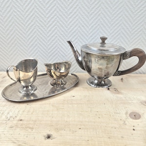 Op de afbeelding: Een zilveren theepot met een houten handvat, een zilveren roomkan en een zilveren suikerpot op een zilveren dienblad.
