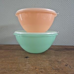 Op de afbeelding: Twee vintage plastic mengkommen, één perzik en één mintgroen, op elkaar gestapeld. Elke kom heeft een bijpassende deksel. De kommen staan op een houten oppervlak.