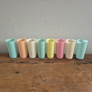 Puede incluir: Colección de siete vasos de plástico de colores pastel. Los colores incluyen azul claro, melocotón, crema, verde menta, amarillo, rosa y blanquecino. Los vasos están colocados sobre una superficie de madera con un fondo gris texturizado.