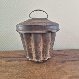 Op de afbeelding: Een vintage metalen cakevorm met een geribbeld ontwerp en een deksel met een handvat. De vorm is roestig en vertoont tekenen van ouderdom.