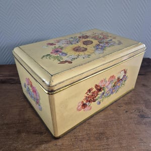 May include: A vintage cream-colored metal box with a floral design. The lid features a bouquet of sunflowers and other flowers in shades of yellow, pink, and purple. The sides of the box have a border of smaller flowers in shades of pink, purple, and red.