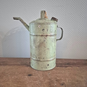 Op de afbeelding: Oude, groen geverfde metalen oliekruik met tuit, handvat en deksel. De cilindervormige kruik heeft horizontale banden en een klein handvat aan de bovenkant. De tuit steekt uit de zijkant en de kruik vertoont gebruikssporen.