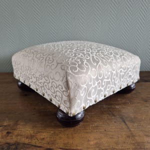 Peut inclure: Un repose-pieds en tissu beige avec un motif en spirale. Le repose-pieds a quatre pieds en bois avec des clous en laiton autour des bords.