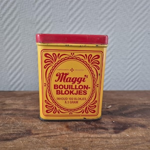 Op de afbeelding: Een vintage gele Maggi bouillonblokjes-blik met een rood deksel. Het blik heeft rode decoratieve krullen en de tekst "Maggi BOUILLON-BLOKJES". Het blik bevat 100 blokjes van elk 3 gram.