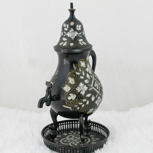 Op de afbeelding: Zwarte metalen theepot met een decoratief bloemenontwerp en een tuit. De theepot heeft drie poten en staat op een bijpassend metalen dienblad.