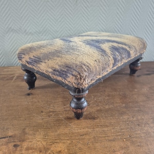 May include: A rectangular footstool featuring a textured, woven fabric top with a dark blue and tan abstract pattern. The base is constructed from dark, stained wood and is supported by four short, turned legs with a rounded, bulbous design.