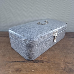 May include: A rectangular, silver-colored metal box with a textured pattern. It features a hinged lid, a latch closure, and a small handle on top. The box is set on a wooden surface.
