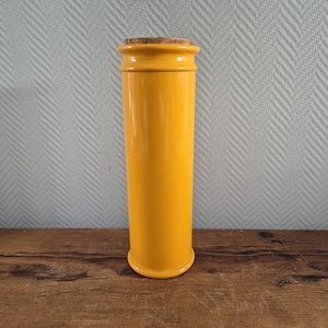 Op de afbeelding: Een hoge, cilindervormige gele pot met een kurken stop. De pot heeft een glanzende afwerking en staat op een houten oppervlak. De achtergrond is een lichtblauwe muur met een subtiel patroon. Deze pot is geschikt voor opslag in de keuken.