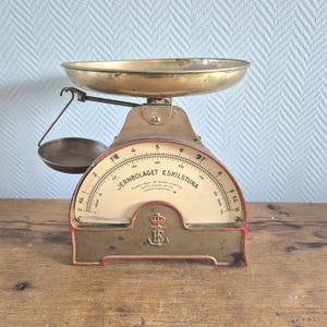 Puede incluir: Balanza vintage de latón y metal con una cara blanca y marcas negras. La balanza dice "Jernbolaget Eskilstuna" y tiene un logotipo de corona y ancla.