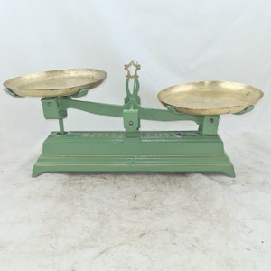 Puede incluir: Una balanza de equilibrio vintage de metal verde con dos platillos de pesaje de latón. La balanza tiene una parte superior en forma de estrella decorativa y una escala graduada en la base.