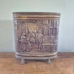 Op de afbeelding: Een rechthoekige metalen container met een gedetailleerd reliëf dat een historische scène uitbeeldt. De container heeft een afgeronde bovenkant en rust op kleine, klauwachtige voetjes. Het reliëf toont figuren in historische kleding tegen een achtergrond van metselwerk.