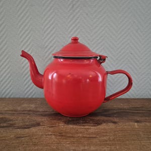 Op de afbeelding: Een vintage rode emaille theepot met een gebogen tuit, een afgeronde body en een bijpassend deksel. De theepot heeft een zwarte rand en een gebogen handvat. Hij staat op een houten oppervlak tegen een lichtgrijze achtergrond.