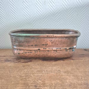 May include: A rectangular copper planter with a weathered patina. The planter has rounded corners and a slightly raised rim. The copper has areas of green oxidation, adding to its antique appearance. It sits on a wooden surface.