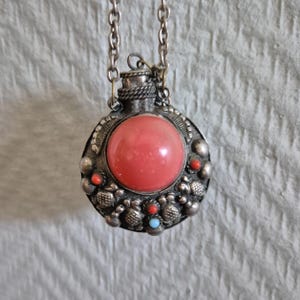 Op de afbeelding: Een zilveren ketting met een ronde hanger, bezet met een roze steen en een zilveren rand. De rand is versierd met kleine zilveren kralen en rode en blauwe stenen.