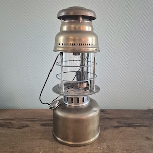 Könnte beinhalten: Eine Messing-Petroleumlampe mit einem Glaszylinder und einem Drahtkäfig. Die Lampe hat einen runden Sockel und einen zylindrischen Tank.