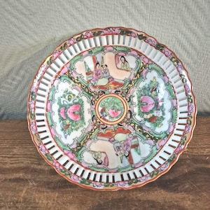 May include: A round, antique porcelain plate with a pierced rim and a scalloped edge. The plate features hand-painted scenes in pink, green, and gold, depicting figures and floral designs. The plate's diameter is approximately 9 inches.