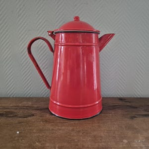 Op de afbeelding: Een vintage rode emaille koffiepot met een deksel, handvat en tuit. De pot is felrood met zwarte randen en staat op een houten oppervlak. Een klassiek keukenartikel.