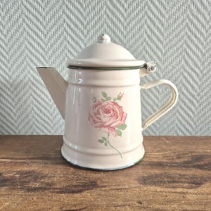 Op de afbeelding: Crèmekleurige emaille koffiepot met deksel en handvat. Een grote roze roos met groene bladeren is op de voorkant geschilderd. De pot heeft een tuit en is afgewerkt met groene en blauwe accenten. De pot staat op een houten oppervlak.