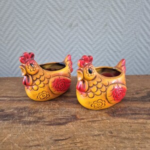 Op de afbeelding: Twee keramische plantenbakken in de vorm van een kip. De plantenbakken zijn geel met rode accenten op de kam, lellen en bloemmotieven. De binnenkant van elke plantenbak is donker. De kippen staan op een houten oppervlak.