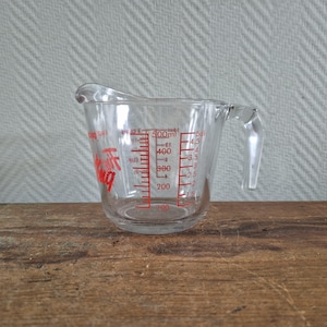 Op de afbeelding: Een doorzichtige glazen maatbeker met een handvat en een schenktuit. De buitenkant is voorzien van rode markeringen die metingen in milliliters tot 500ml en deciliters tot 5dl aangeven, samen met kopjes-eenheden en de tekst Pyrex.