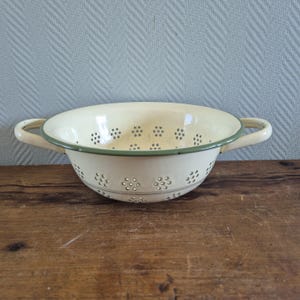 May include: Cream enamel colander with a green rim and handles. The kitchen utensil has small, evenly spaced holes for draining liquids. The colander is set on a wooden surface, a classic piece of kitchenware.