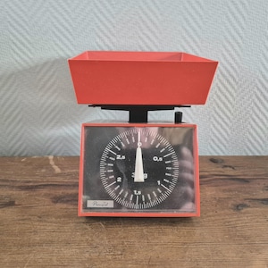 May include: Red kitchen scale with a black dial and a red bowl. The dial has markings for grams and ounces. The scale is labelled "Rowenta".