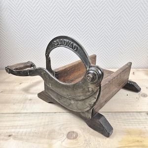Könnte beinhalten: Ein antiker Brotschneider mit Holzsockel und gebogener Metallklinge. Auf der Metallklinge ist das Wort "RAADVIAD" eingeprägt. Der Griff ist aus Holz und an der Metallklinge befestigt. Der Schneider ist zum Schneiden von Brot bestimmt.