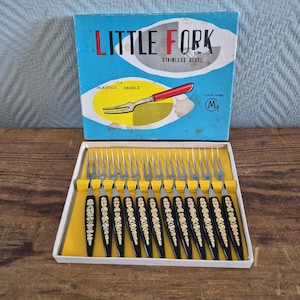 Op de afbeelding: Vintage set van roestvrijstalen vorken met zwarte handvatten en gouden bloemmotieven, gepresenteerd in een doos met het opschrift "LITTLE FORK". De doos is blauw en wit met een rode en gele vorkafbeelding. De vorken liggen in een gele tray.