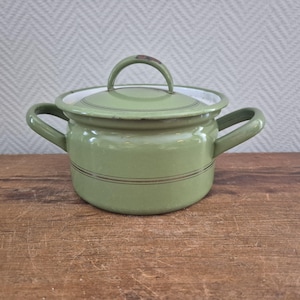 Op de afbeelding: Een vintage, lichtgroene emaille kookpot met een deksel en twee handvatten aan de zijkant. De pot heeft een witte rand en twee dunne, donkerbruine strepen rond de body. Het deksel heeft een lusgreep en een klein, vervaagd ontwerp. De pot staat op een houten oppervlak.