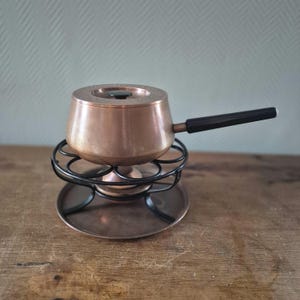 Puede incluir: Una olla de fondue de cobre con un mango negro se asienta sobre un soporte de metal negro con una base de cobre. La olla tiene una tapa y está diseñada para derretir chocolate o queso.