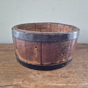Op de afbeelding: Een rustieke houten emmer met donkere metalen banden. De emmer is gemaakt van verticale houten planken die bij elkaar worden gehouden door twee donkere metalen banden. Het hout heeft een warme, bruine tint en de metalen banden zijn donkergrijs. De emmer staat op een houten oppervlak.