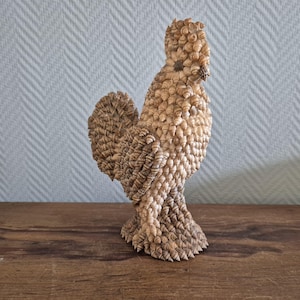 Puede incluir: Una figura de gallo hecha completamente de conchas marinas. El gallo está de pie sobre una superficie de madera. El gallo es marrón y blanco.