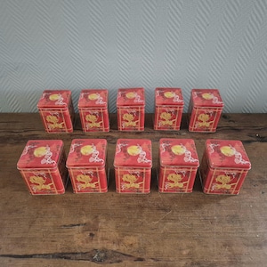 Op de afbeelding: Tien vierkante blikken doosjes in rood met gouden en rode ontwerpen. Elk doosje heeft een drakenontwerp met een gele cirkel in het midden. De doosjes zijn gerangschikt in twee rijen van vijf.