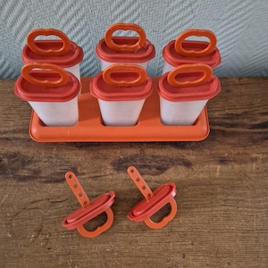 Peut inclure: Un ensemble de moules à sucettes glacées en plastique orange, comprenant six moules individuels avec un couvercle et une poignée rouges. Deux poignées rouges supplémentaires sont devant l'ensemble. L'ensemble est sur une surface en bois.