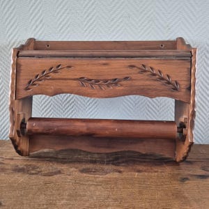 May include: A vintage wooden paper towel holder with a shelf. The holder is made of dark brown wood and features carved leaf designs. The bottom has a wooden dowel for holding the paper towels. The shelf is scalloped.