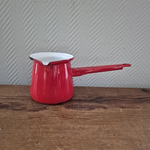 Puede incluir: Una pequeña cafetera roja de esmalte con un mango largo. La cafetera tiene un interior blanco y un borde negro. La cafetera está sobre una superficie de madera.