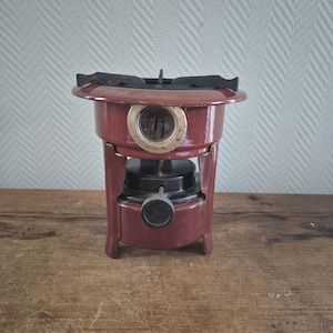 Op de afbeelding: Een vintage rode campingkachel met een zwarte brander en een knop om de vlam te regelen. De kachel heeft een ronde basis en drie poten.