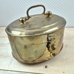 Op de afbeelding: Een vintage ovale doos van messing met een scharnierend deksel en een slot. De doos heeft een handvat aan de bovenkant en is versierd met een eenvoudig ontwerp.