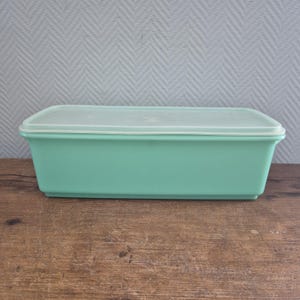 Op de afbeelding: Een rechthoekige, vintage, turquoise plastic container met een doorschijnend deksel. De container staat op een houten oppervlak. Het deksel heeft een subtiel, reliëf ontwerp. De container is waarschijnlijk voor voedselopslag.