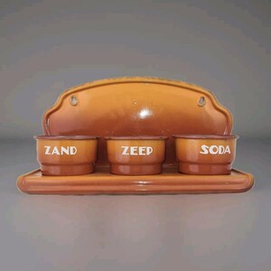 Könnte beinhalten: Ein vintage orangefarbenes Email-Wandregal mit drei kleinen Behältern mit den Aufschriften "ZAND", "ZEEP" und "SODA".