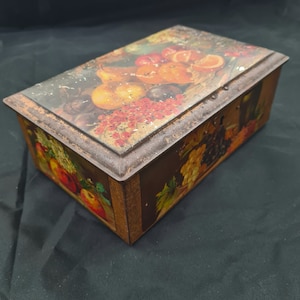 Op de afbeelding: Een vintage blikken doos met een kleurrijke illustratie van fruit, waaronder sinaasappels, druiven en peren. De doos heeft een versleten, rustieke uitstraling en is perfect voor het opbergen van kleine voorwerpen.