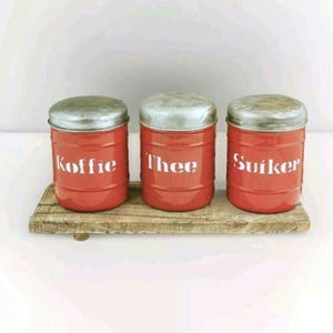 May include: Three red metal canisters with silver lids, labeled "Koffie", "Thee", and "Suiker" on a wooden shelf.