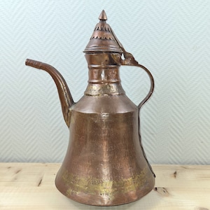 Op de afbeelding: Een koperen theepot met een decoratief deksel en een tuit. De theepot heeft een gehamerde afwerking en een gouden band rond de bodem.