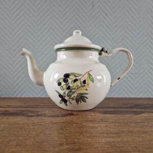 Op de afbeelding: Crème-kleurige emaille theepot met een deksel en handvat. De theepot heeft een groene rand en een geschilderd olijftakontwerp met groene bladeren en zwarte olijven. De theepot staat op een houten oppervlak.